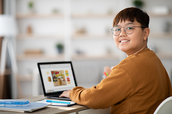 Cheerful chubby chinese kid with glasses sitting at desk at home, using laptop with empty screen, having video conference with teacher or tutor, surfing on Internet, mockup, copy space